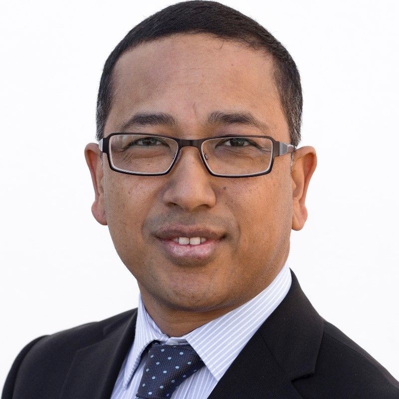 Man in glasses wearing a suit and tie with a neutral expression against a white background.